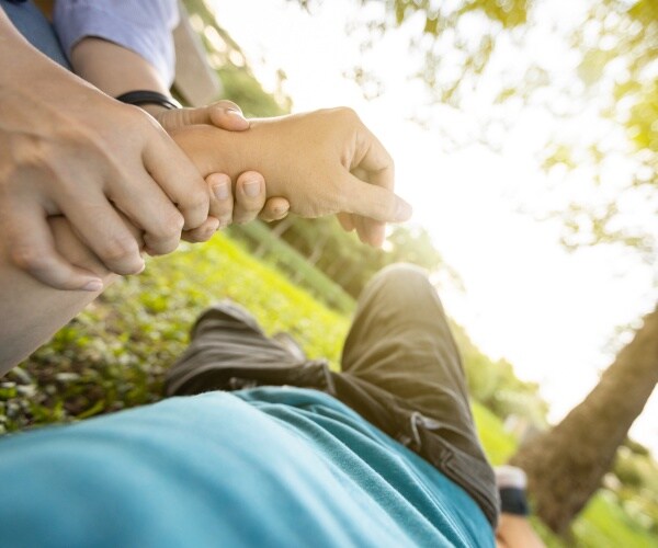 man sick on ground outside on hot day with a woman checking his pulse