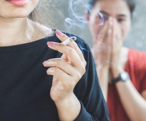 woman smoking a cigarette with another woman behind her getting smoke in her face