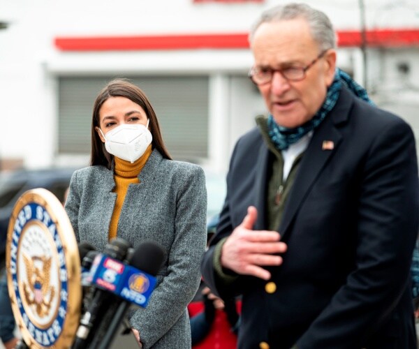 standing outdoors, aoc listens to chuck schumer speak at a podium