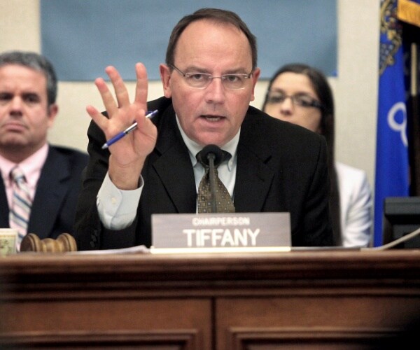 tom tiffany holding up four fingers and a pen in his hand while speaking at a hearing