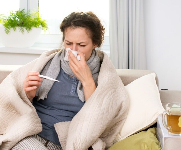 woman sick on couch, blowing her nose, taking her temperature