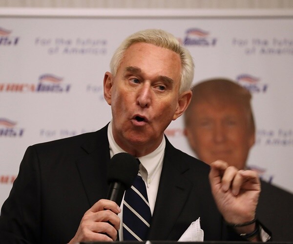 roger stone stands in front of a photo of president donald trulmp