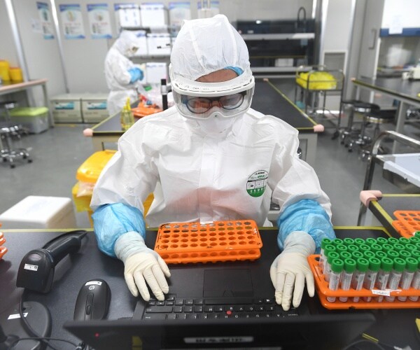 lab researcher typing on a laptop in a white protective suit and face mask with sample tubes next to her