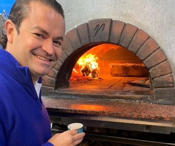 a man stands and smiles with a brick oven flaming grilling pizza in the background