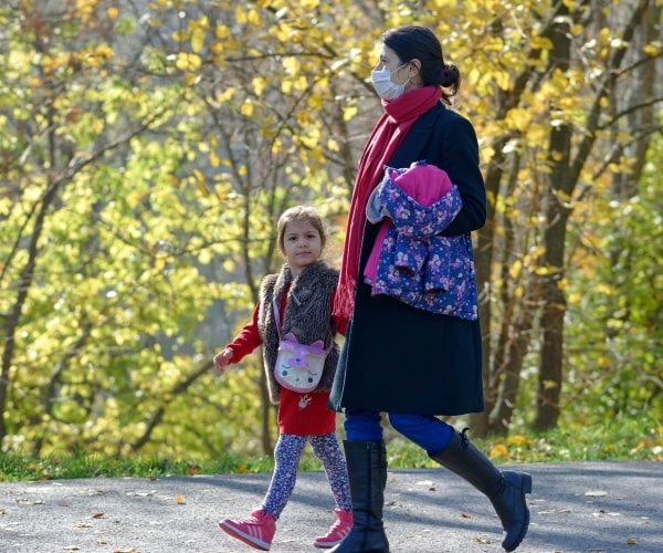 A mother wearing a surgical mask walking in park with daughter looking at camera without mask