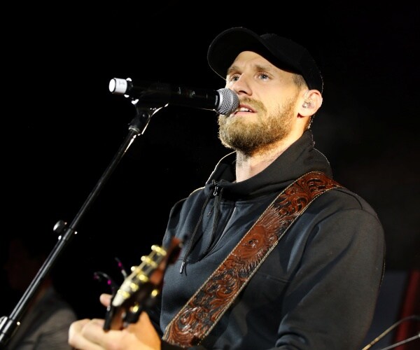 rice in a black jacket playing a guitar and singing into a mic