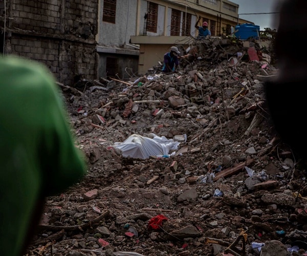 body under white sheet in rubble of collapsed building