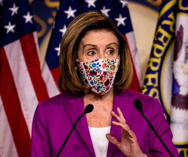 wearing a mask, nancy pelosi speaks in front of flags