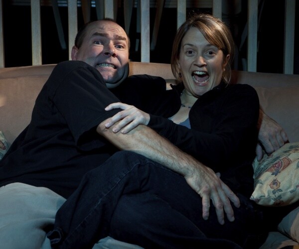 man and woman on couch, grabbing each other and looking scared while watching scary movie