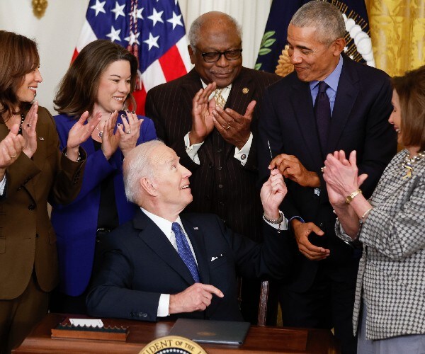 biden seated at a desk handing a pen to obam with kamala harris nancy pelosi and legislators standing behind him