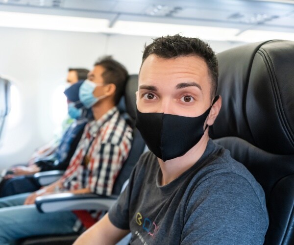 man on airplane with mask on looking at camera, others around him also wearing masks