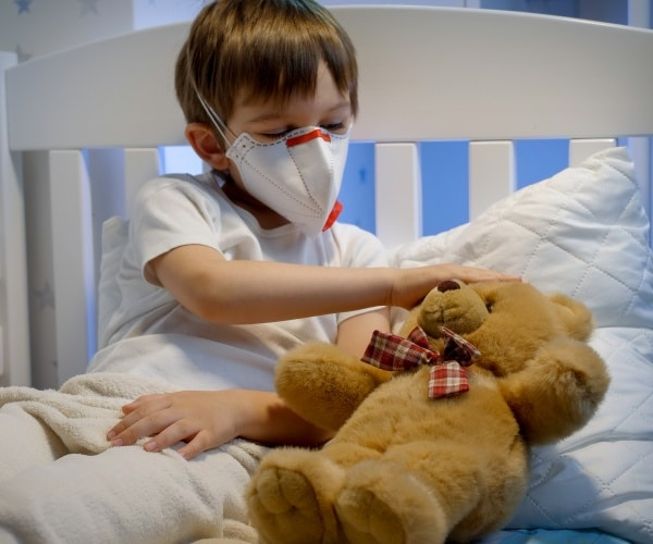 boy sick in bed with teddy bear -- both wearing masks