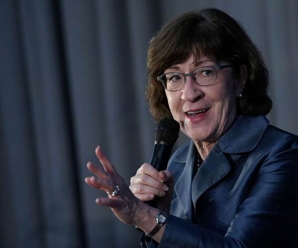 susan collins wearing glasses in a dark blue coat speaking into a mic with her hand out