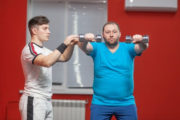 A heavy man lifting weights with a trainer in a gym