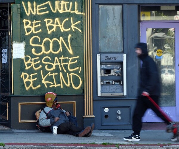 homeless man wears a jacket and leans against a boarded up shop