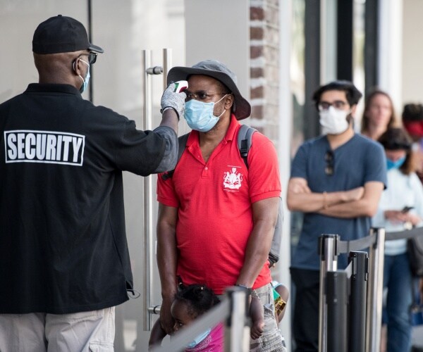 people wearing masks line up outside a store and a security guard takes the temperature of a man