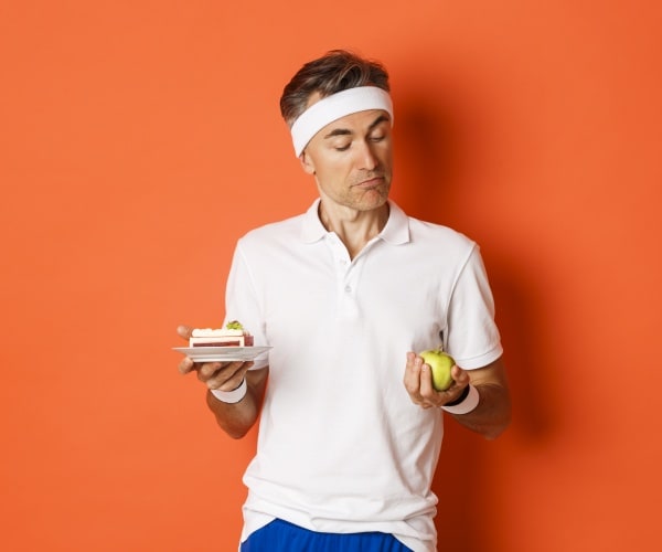 man in workout clothes and headband looking at apple in one hand with cupcake in other