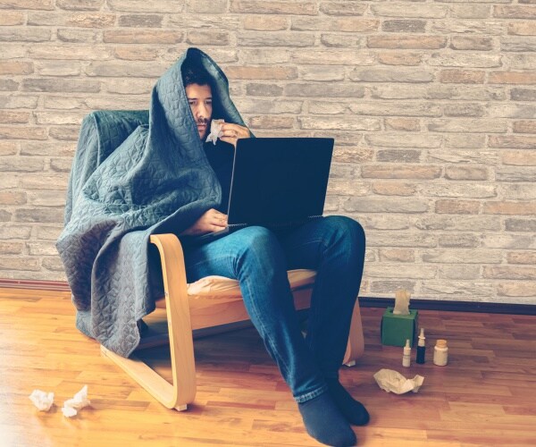 man sick in chair with blanket over head looking at laptop with tissues and medicines around him on floor