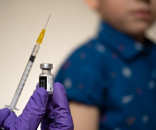 gloved hand holding up COVID vaccine vial/syringe and child in background