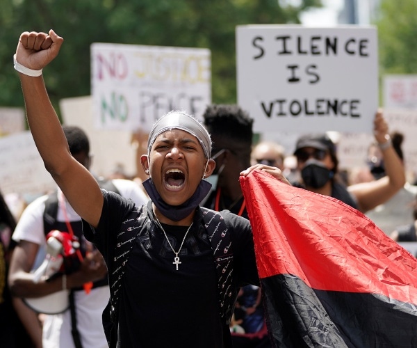 young black man in a black tshirt holds a fist up with people holding up signs in the background