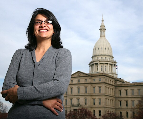 rashida tlaib stands smiling with her arms folded and the michigan capitol building in the background