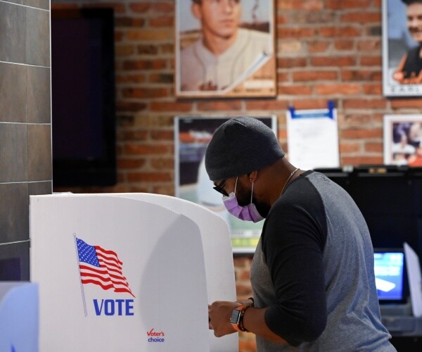 man wearing a gray shirt and purple face mask votes at a booth with a white privacy divider