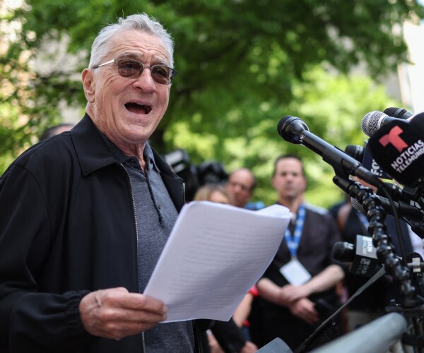 robert de niro reads from a piece of paper while standing in front of multiple press microphones