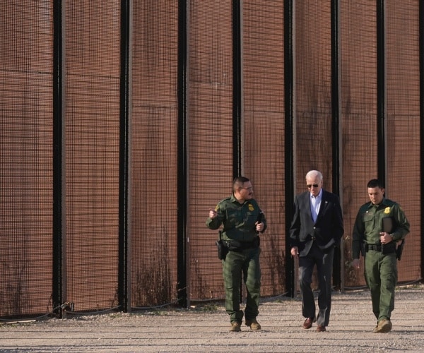 joe biden walks with border patrol agents on left and right with wall in background