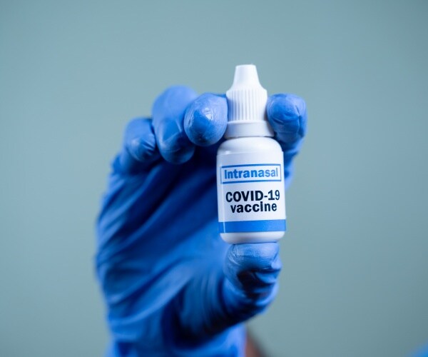 hand with blue surgical glove holding up generic spray labeled "COVID-19 vaccine"