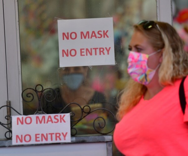 woman in a bright neon top and mask walks by window with a sign saying "no mask no entry"