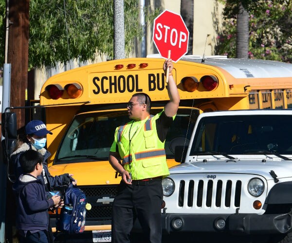 a woman wearing a blue face mask walks with her son as a crossing guard directs traffic