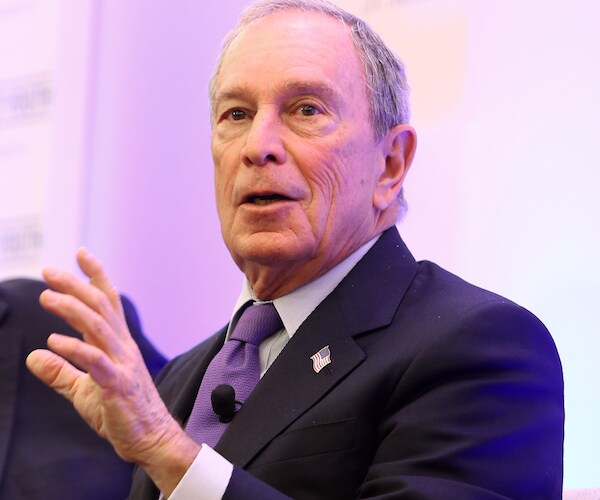 michael bloomberg raises his left hand as he looks left and delivers comments during a conference