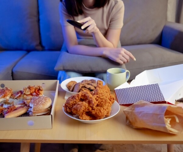 woman sitting on couch with tv remote and pizza, hamburgers, fries, other unhealthy foods in front of her