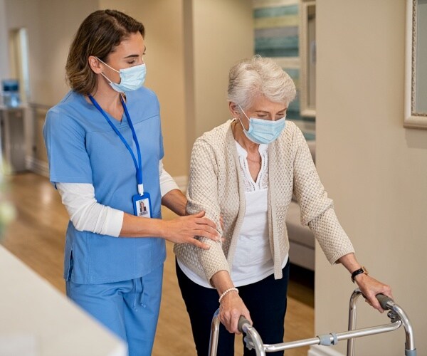 nurse and nursing home resident with walker and both wearing masks walking down hall