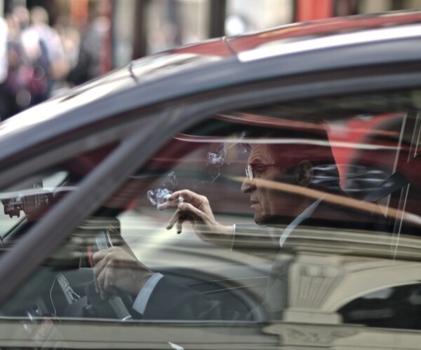 older man in suit, driving car in city, smoking