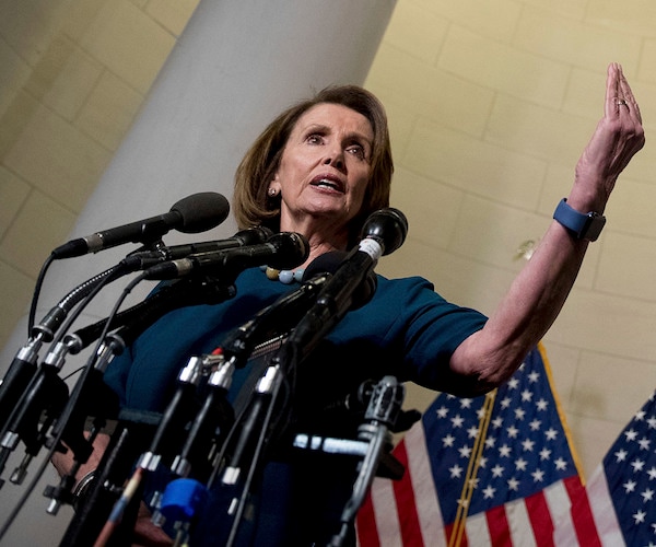 nancy pelosi, speaking into a gaggle of mics, raises his left hand, palm and fingers up, high into the air