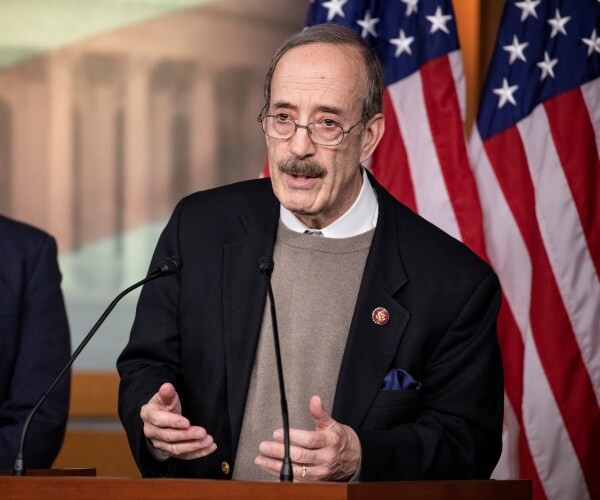 eliot engel in a black jacket and tan/gray sweater speaking in front of two american flags