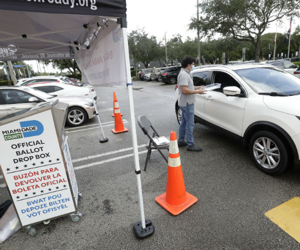 A poll worker in shirt, jeans and mask waits as a voter drops off their Vote-by-Mail ballot