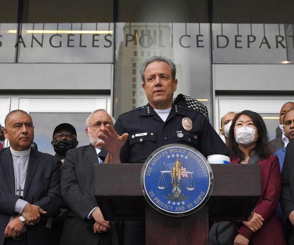 michel moore speaks at a lapd lectern with faith leaders behind him, some wearing masks.