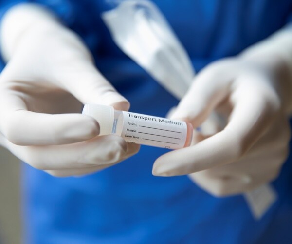 nurse in a blue protective suit and white gloves holds a small tube labeled transport medium