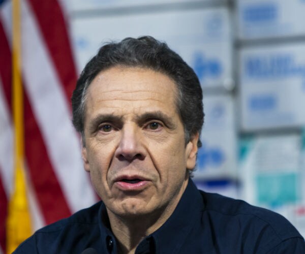 ny gov. andrew cuomo is shown in a blue button down shirt with the us flag in the background