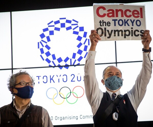 protester holds sign that says cancel the tokyo olympics in front of tokyo olympic logo