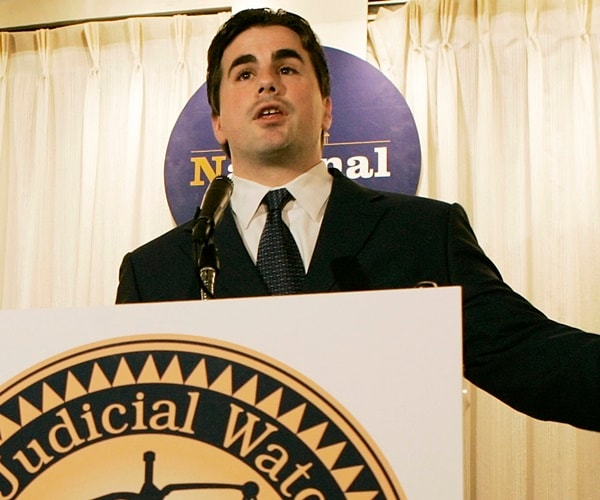 tom fitton speaksat a podium with a judicial watch sign on it