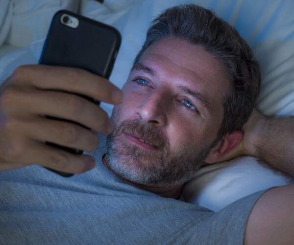 man lying in bed in dark looking at smartphone, with light from screen on his face