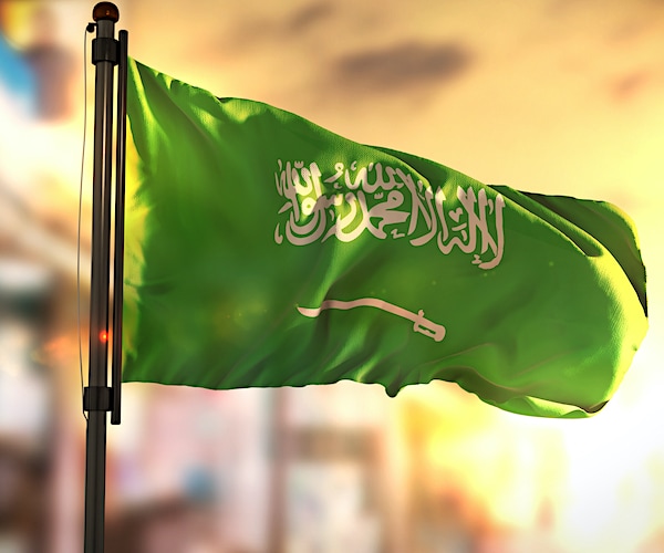 Saudi Arabia Flag Against City Blurred Background At Sunrise Backlight Sky