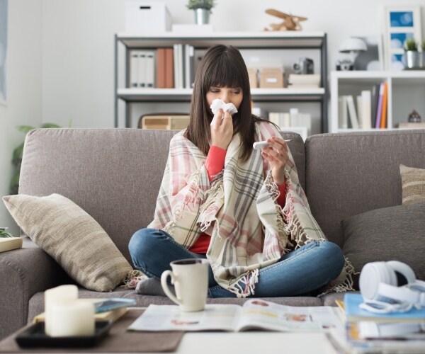 woman sitting on couch not feeling well with blanket, tea, tissues, taking temperature