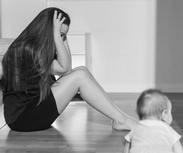 woman sitting on floor with head in her hands, her baby crawling near her