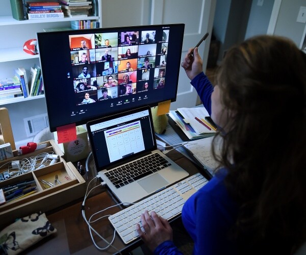 teacher wearing a blue top sits in front of a laptop and a desktop computer on a zoom call with a class