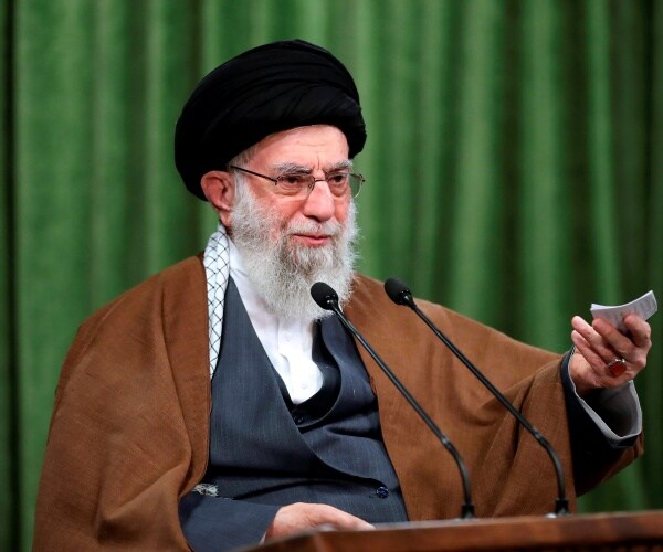 khamenei in a brown shawl and black turban sitting in front of a green curtain