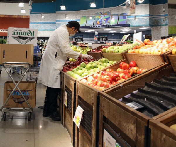 a grocery story worker restocks fruit while wearing gloves, masks and a protective work jacket